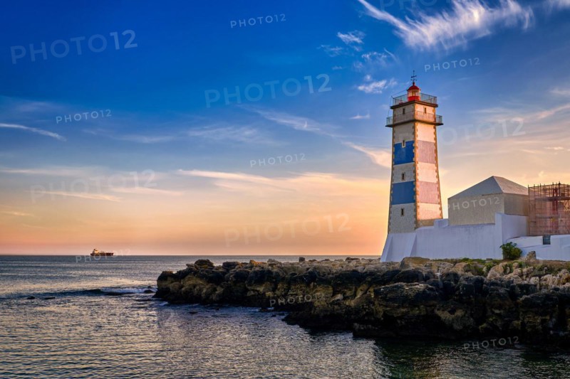 Dramatic sunset on sea shore and Santa Marta lighthouse in Cascais, Portugal. Colorful sky, clouds, sunlight, low sun, calm ocean waters, local landmark, safety and navigation tower, rocky shore