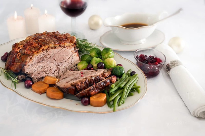 Roast pork ham with crust, green beans, Brussel sprouts, carrots, cranberry and sauce on white table cloth with candles and Christmas decoration, festive dinner, selected focus, narrow depth of field