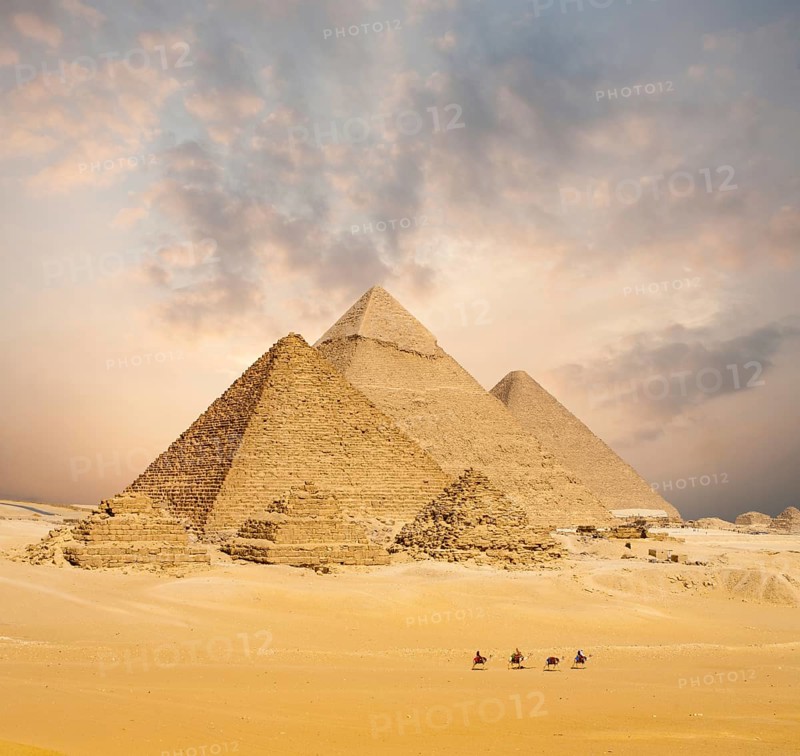 Dramatic sunset behind all distant Egyptian Pyramids together with line of camels walking in foreground in Giza, Cairo, Egypt. Wide compressed telephoto shot with plenty of copy space