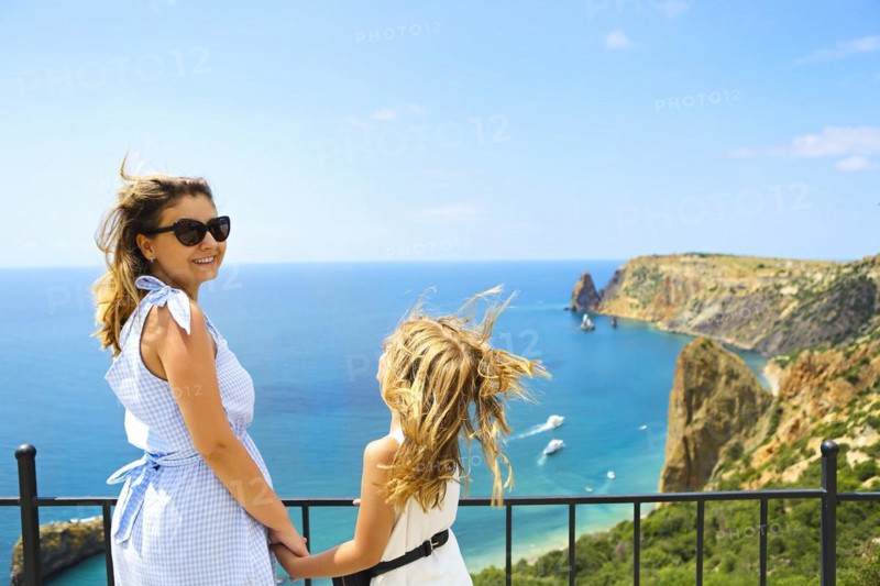 Mother and little girl standing next to fence on viewpoint and admiring picturesque view of rocky sea coast in sunny day with clear sky and blue water