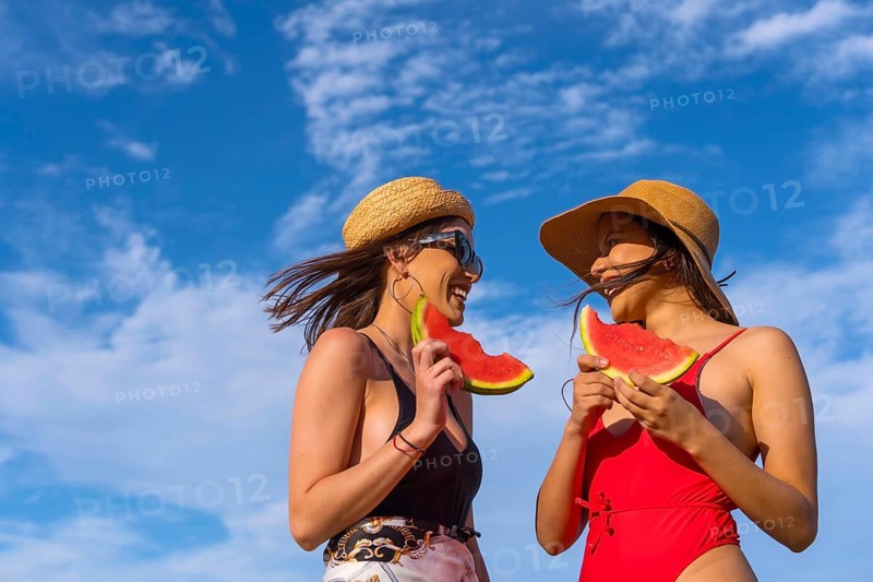 Female friends at the beach in summer eating a watermelon with the sky in the background, shot from bottom to top