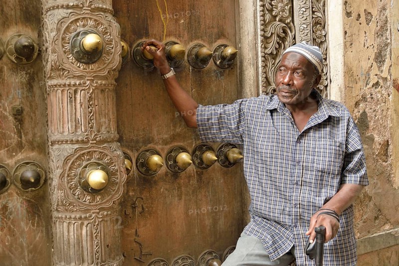 Elderly man leaning against a wooden door in the historic town centre, Zanzibar City, Zanzibar, Tanzania, Africa