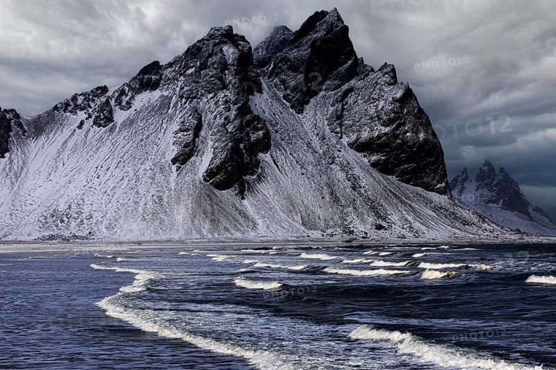 White foamy waves on the beach of Stokkness, behind snowy rocky slopes of Vestrahorn, near Höfn, Sudausturland, Iceland, Europe