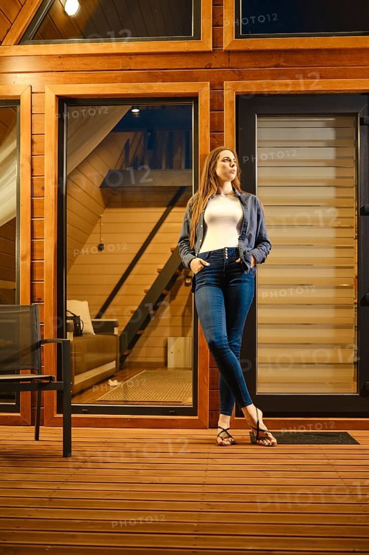 A beautiful brunette woman stands on the doorway of a large wooden cabin in the evening