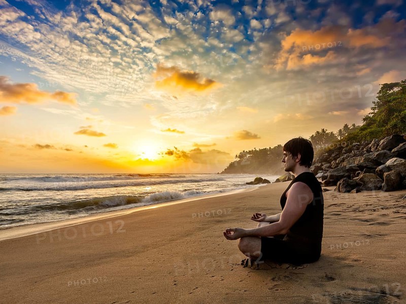 Young sporty fit man doing yoga meditating in padmasana lotus pose on tropical beach on sunset