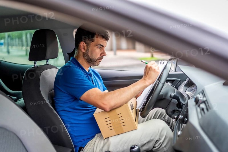 Side view from inside a vehicle of a serious delivery man writing and inventorying packages on a car