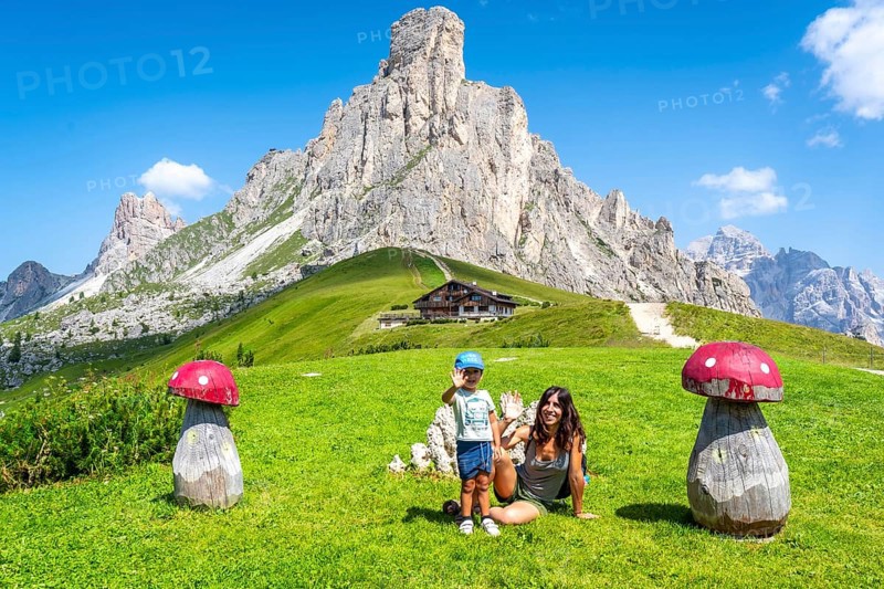 Mother and son waving near big wooden mushrooms with scenic view of nuvolau peak and mountain hut at giau pass in dolomites