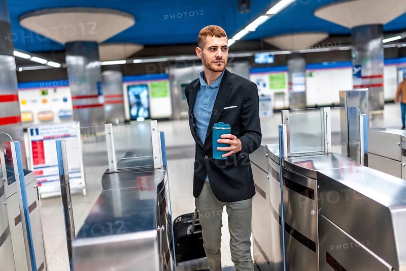 Young businessman going to work passing through subway turnstile holding reusable coffee cup and pulling trolley suitcase
