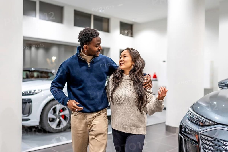 Happy multiethnic couple walking and choosing an electric car in a dealership, hugging and smiling