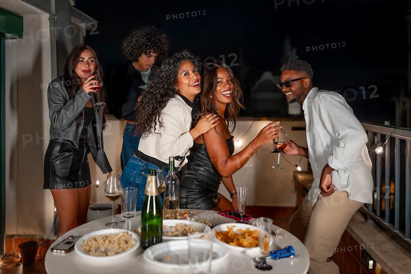 Playful drunk multi-ethnic friends drinking champagne and dancing on roof at night