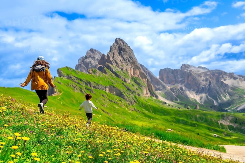 Tourists running through a field of wildflowers with the seceda peaks in the dolomites mountain range of italy in the background on a beautiful summer day