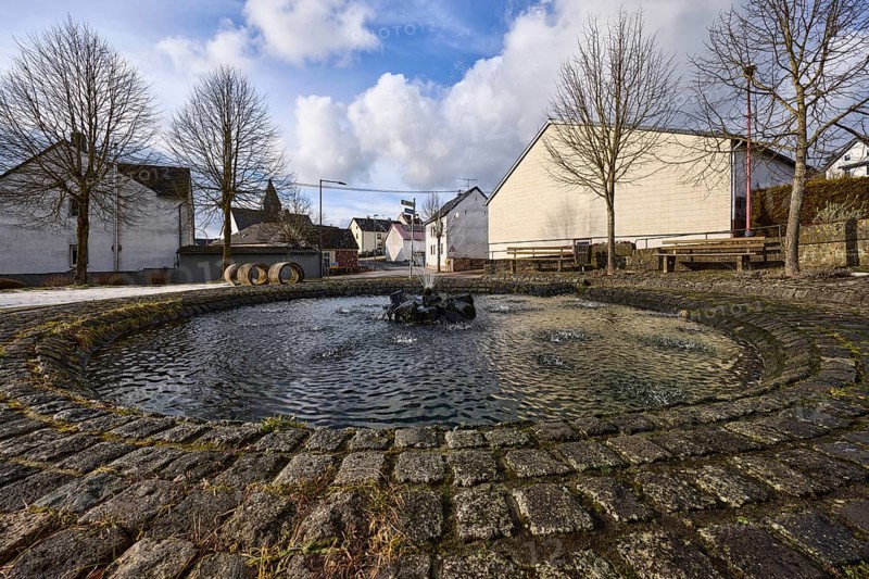 Fountain, water fountains, fountain, houses, trees, blue sky with cumulus clouds, frog perspective, intersection Im Höfchen, Hauptstraße and Heyrother Straße, Dreis-Brück, Eifel, Vulkaneifel district, Rhineland-Palatinate, Germany, Europe