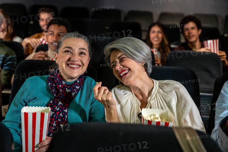 Two cheerful senior women laughing and eating popcorn while watching a movie at the cinema, sharing a joyful moment together