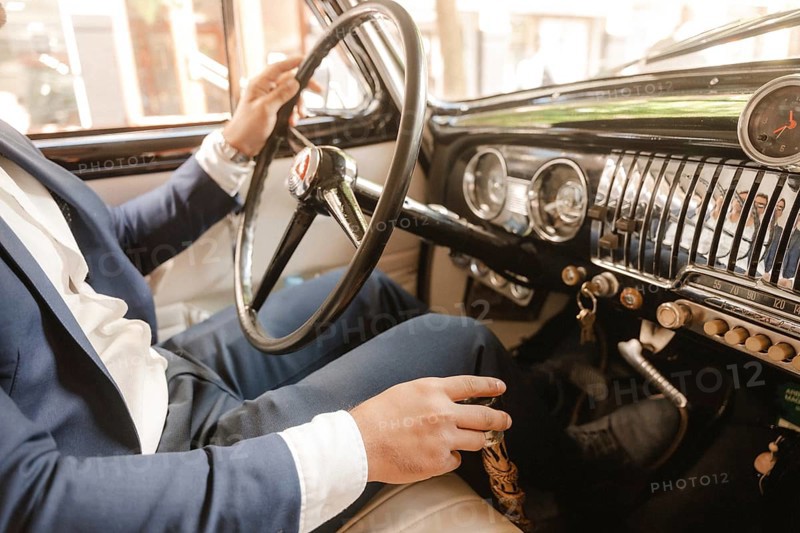 A man in a suit is driving a car with a black steering wheel. The car is a vintage model and the man is wearing a blue suit. Concept of elegance and sophistication