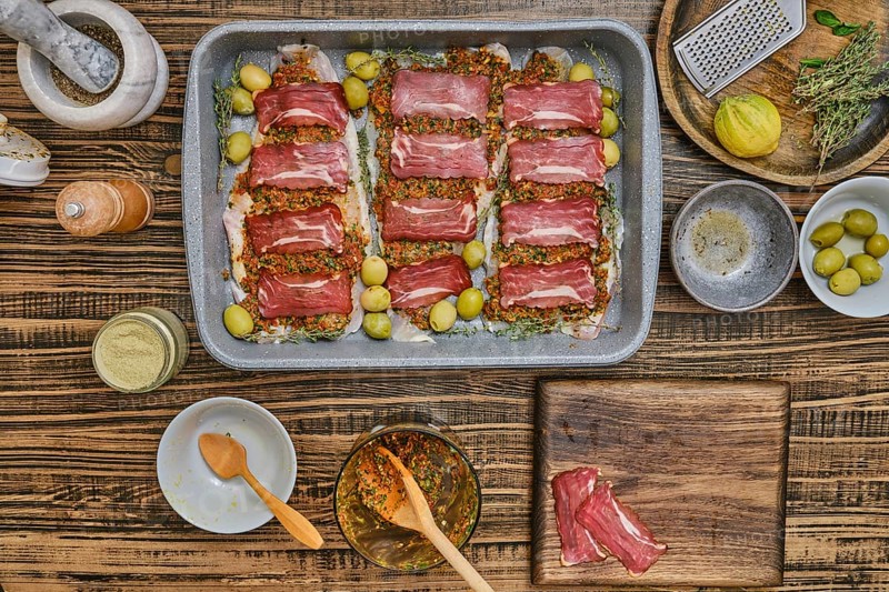 Fresh fish fillets layered with bacon and seasoning, herbs, and olives sits on a baking tray. Surrounding ingredients include spices and utensils, showcasing the artistry of cooking in a home kitchen