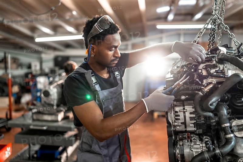 Young mechanic using pliers, repairing a car engine hanging from a chain hoist, inside a garage
