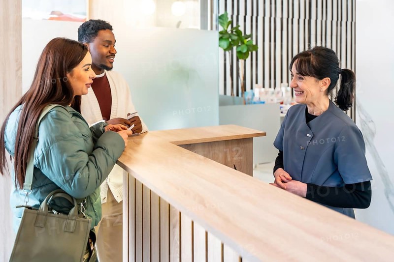 Interracial couple filling out paperwork at the reception desk of a modern dental clinic, interacting with the friendly receptionist