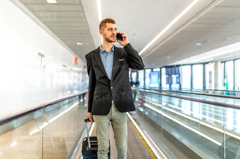 Businessman pulling suitcase and talking on smartphone while walking on moving walkway in airport