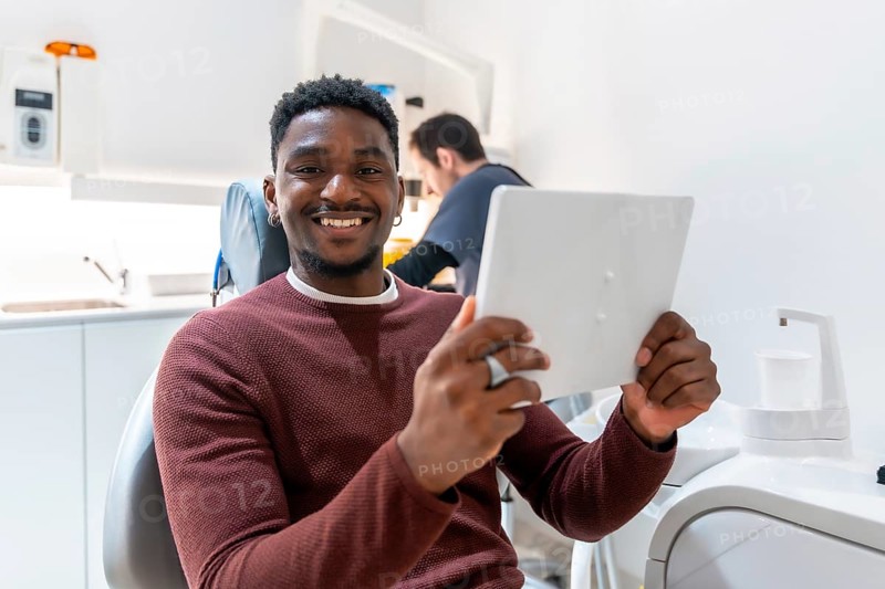 Smiling patient using digital tablet, possibly reviewing treatment plan or x rays, while dentist works in background of modern dental clinic