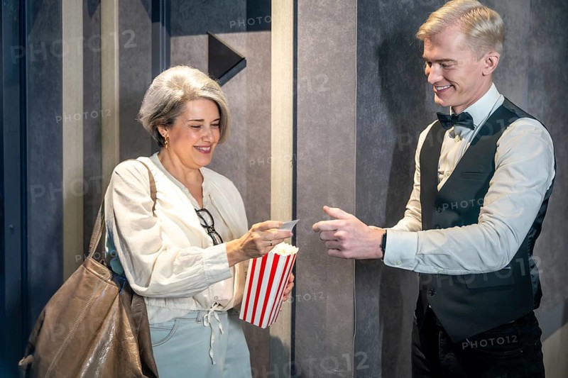 Elegant mature woman handing ticket to smiling usher while holding popcorn bucket, ready to enjoy movie session