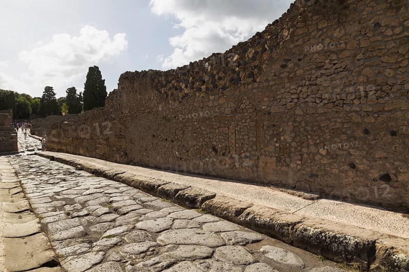 Long narrow stone sidewalk and street made of large grey smooth and worn lava stones plus remnant of old stone and terracotta brick wall structure at the ancient ruins of Pompeii, Campania region, Italy, Europe
