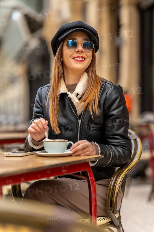 Young woman wearing sunglasses and baker boy hat stirring cappuccino at outdoor cafe table with smartphone nearby