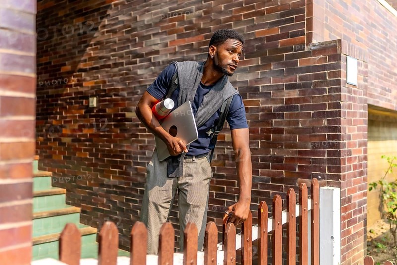 Young african businessman leaving a house in a residential district to go to work holding a laptop and a coffee thermos