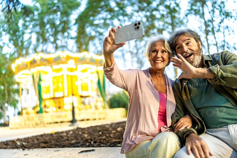 Senior couple waving while taking selfie sitting on bench in the city fair