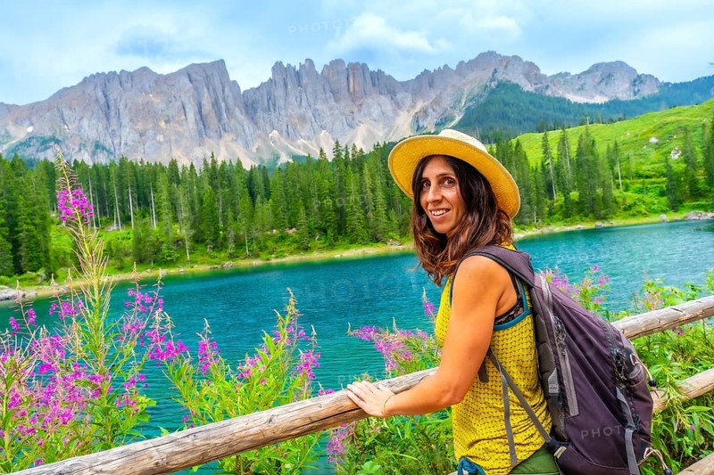 Female tourist with backpack and straw hat enjoying the landscape of lake carezza and latemar mountain in the dolomites, italian alps
