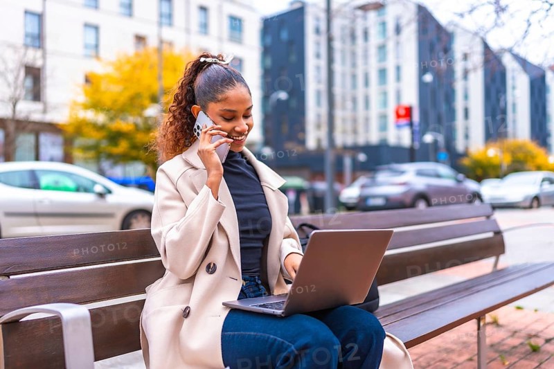 African businesswoman talking to the phone and using laptop sitting on a city bench