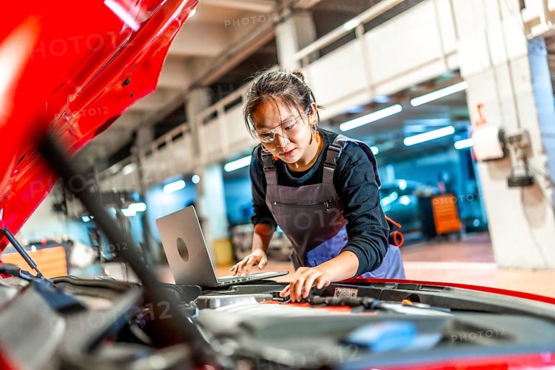 Young woman working as a mechanic, using a laptop while inspecting the engine of a red car in a busy garage workshop