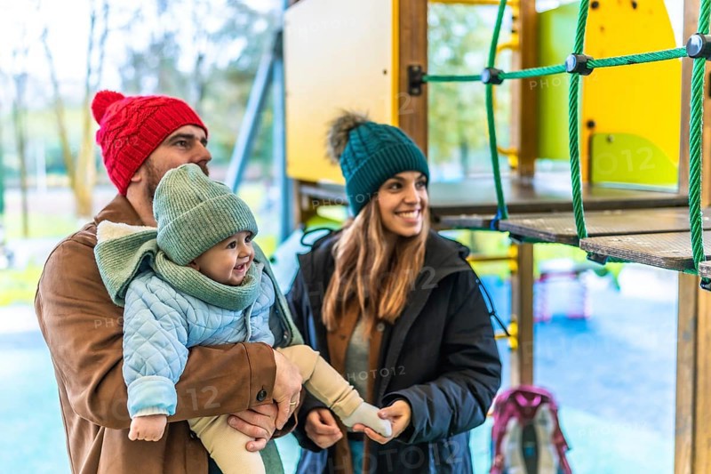 Smiling parents holding their baby at the playground, wearing winter clothes, enjoying a family moment