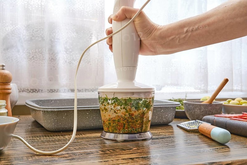 A hand blending device is being used to process fresh herbs and vegetables in a well-organized kitchen. Sunlight filters through a window, creating a warm atmosphere for cooking