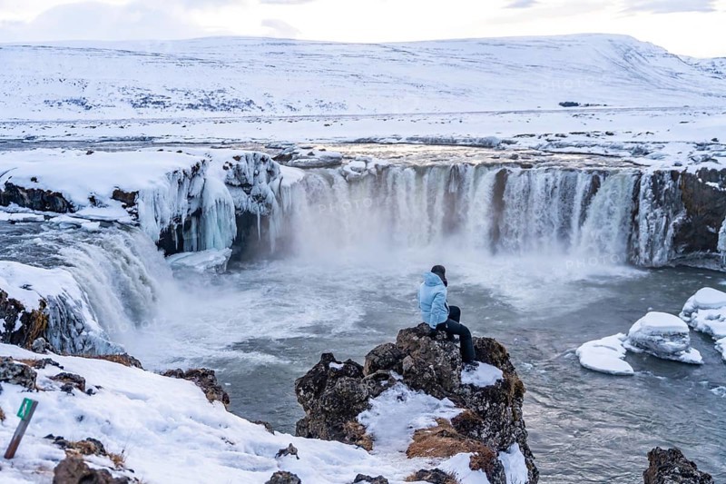 Woman hiker sitting looking at frozen and snowy Godafoss waterfall in winter in Iceland