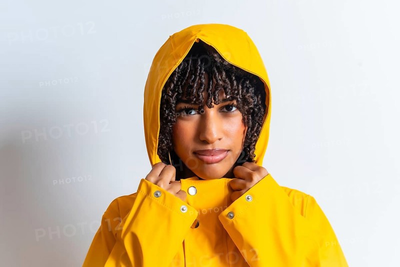 Studio portrait of a young woman with curly hair wearing a yellow raincoat and pulling the hood strings