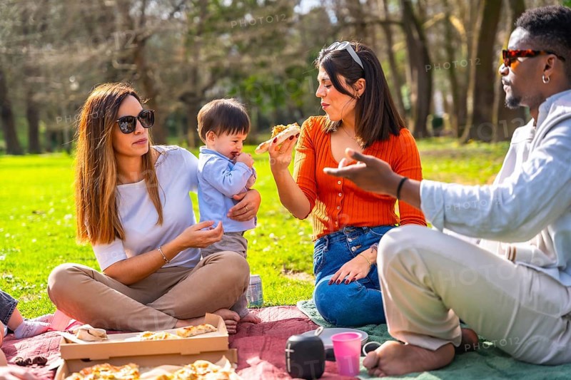 Family and friends enjoying pizza while picnicking in a vibrant green park, laughing and sharing joyful moments under the sunny sky