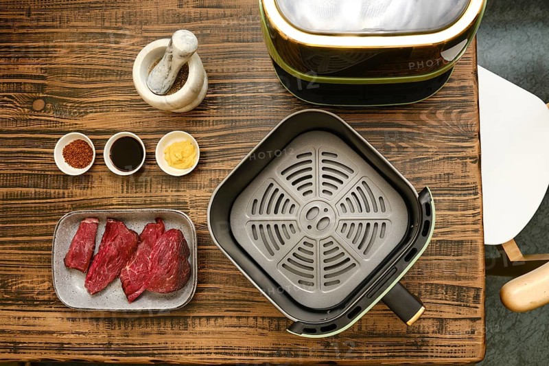 Fresh cuts of meat are arranged alongside several bowls of spices and sauces on a wooden table. A cooking appliance sits nearby, ready for a grilling or air-frying session
