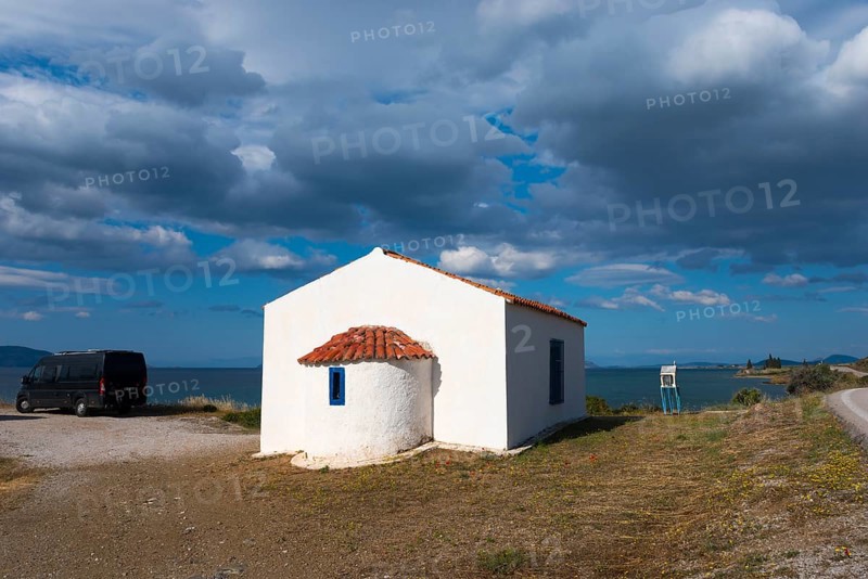 White building with a red roof by the sea, surrounded by a cloudy sky and peaceful countryside. A motorhome is parked next to it. Chapel near Vlacheika, Vlachaiika, Attica, Peloponnese, Greece, Europe