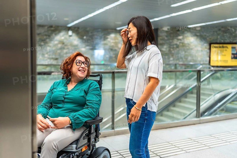 Smiling caucasian adult disabled woman and asian female friend talking waiting for subway elevator