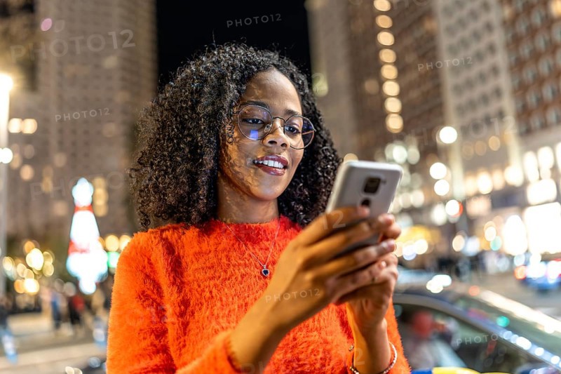 Young woman wearing an orange sweater, smiling while using a smartphone on a bustling city street, surrounded by festive holiday lights at night