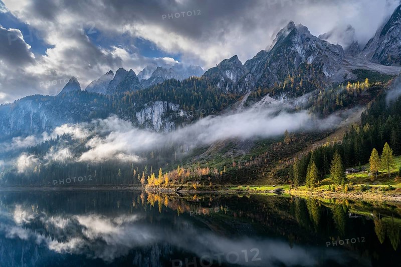The Vordere Gosausee in autumn with a view of the Gosaukamm. Cloudy sky with sun. Trees in autumn leaves. Reflection. Overcast with clouds. Vorderer Gosausee, Gosau, Gosau valley, Salzkammergut, Upper Austria, Austria, Europe