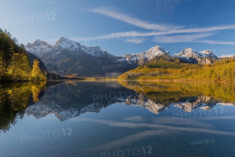 Lake Almsee in autumn with a forest and mountains reflected in the lake. A slowly dissipating fog moves over the lake. Blue sky