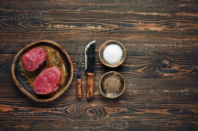 Two raw steaks on a wooden plate alongside bowls of salt and pepper, with old forged knife and fork placed next to them, a cooking session or barbecue preparation