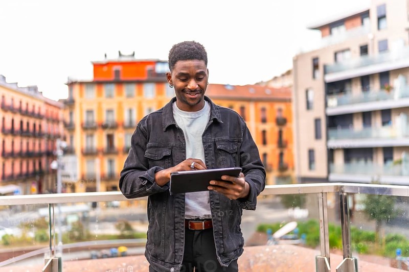 Frontal three quarter length photo of a happy young african man using digital tablet standing in a city