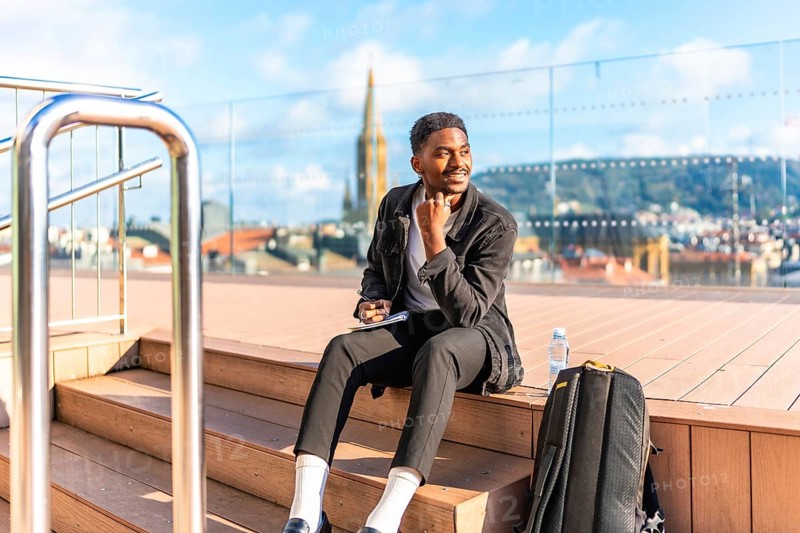 Happy african man sketching by hand in a notebook sitting on a rooftop with city views