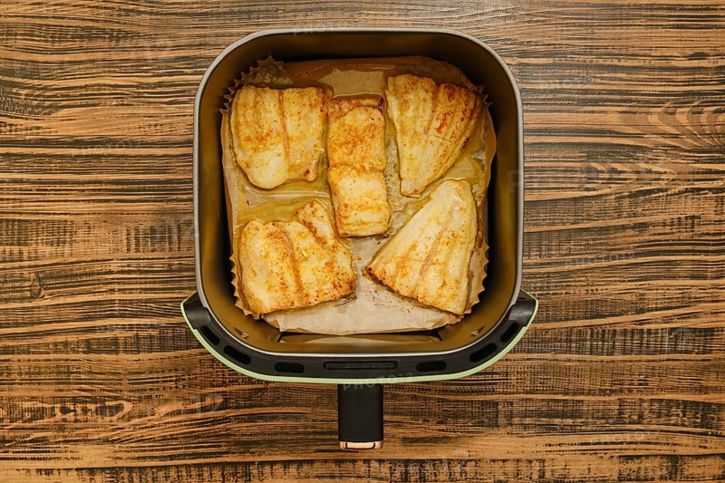 Five golden brown grilled fish fillets are neatly placed inside an air fryer basket. The background features a rustic wooden table, showcasing a cozy cooking environment