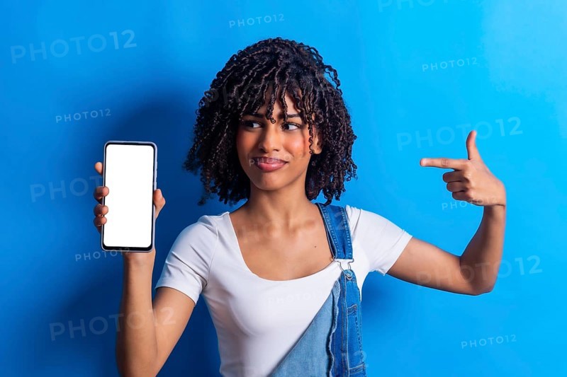 Studio portrait of a young woman holding a smartphone with blank white screen and pointing at it with index finger, isolated on a blue background