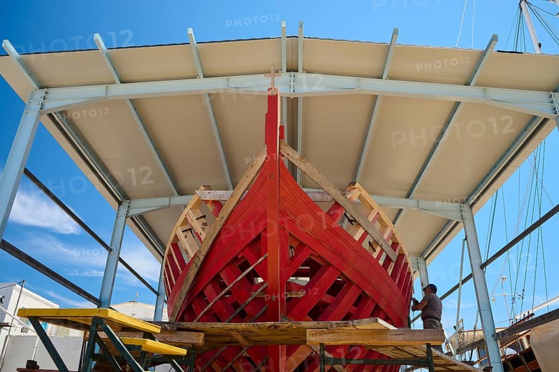 Front view of a partially built red wooden ship in a covered shipyard under a clear sky, Patmos Marine, boatyard, Diakofti, Patmos, Dodecanese, Greek Islands, Greece, Europe