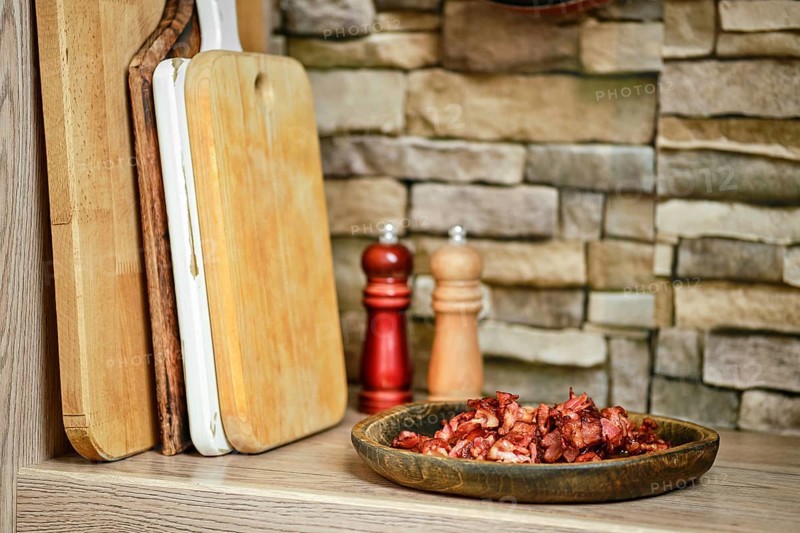 A rustic kitchen features a plate of chopped bacon next to wooden cutting boards. Two shakers, one wooden and one red, sit in the background, adding to the warm atmosphere