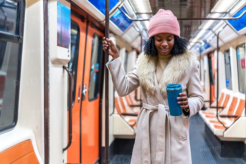 Smiling young black woman holding reusable coffee cup while standing on moving subway train